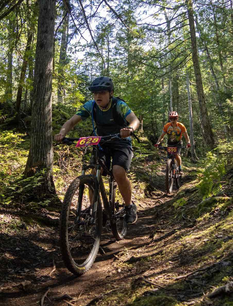 Two riders race down a forested trail